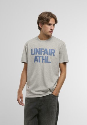 Harmaa puuvillapaita, jossa on rohkeaa sinistä tekstiä, joka kuuluu "UNFAIR ATHL." Lyhyet hihat, pyöreä kaula-aukko, rento malli, sileä pinta.