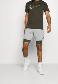 Camiseta Nike verde oliva con logo de camuflaje, combinada con pantalones cortos grises con ribete verde y logo negro de Nike. Zapatillas blancas completan el look.