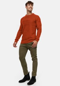 Suéter de punto naranja con un diseño texturizado, de manga larga y con puños de canalé, combinado con pantalones oliva ajustados y botines negros.