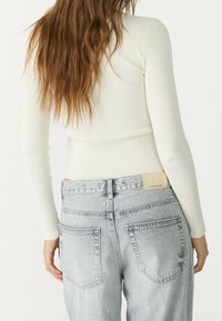White ribbed long-sleeve top paired with light gray high-waisted jeans. Jeans feature a branded tag at the back and two rear pockets.