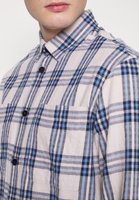 Napapaita kiinni napilla, jossa on ruudullinen kuvio sinisessä ja harmaassa. Benäys kaulus, etutasku ja tummat napit huokoisessa kankaassa.