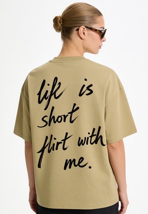 Frau mit Dutt trägt eine Sonnenbrille und ein beigefarbenes Shirt mit schwarzem Text mit der Aufschrift "life is short flirt with me" auf dem Rücken.