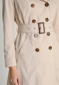Gabardina beige con cintura ceñida, diseño de doble botonadura, grandes botones marrones y bolsillos laterales. Tejido suave con un ajuste entallado.