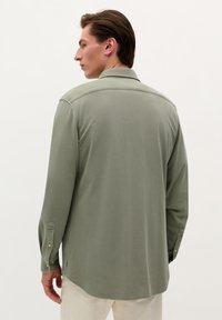 Cămașă cu mâneci lungi, de culoare verde măslin, cu guler și mansete cu nasturi, precum și slituri laterale. Material texturat cu detalii de cusătură subtile.