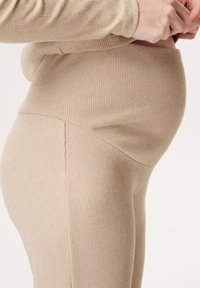 Beżowe, prążkowane legginsy ciążowe z elastycznym paskiem w talii, bezszwowym wykończeniem i rozciągliwym materiałem dla komfortu, pokazane z boku.