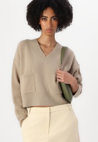 Pulover bej din tricot cu decolteu în V, două buzunare frontale și textură rasucită, asortat cu pantaloni bej deschis și o geantă verde de umăr.