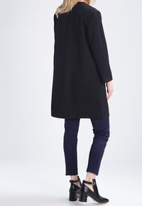 Černý dlouhý kabát z pevné látky, kombinovaný s tmavě modrými úzkými kalhotami. Obuv tvoří černé kožené kotníkové boty na podpatku s přezkou.