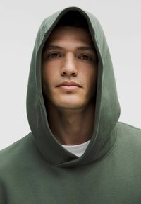 Joven con barba clara que lleva un sudadera con capucha verde, mirando directamente a la cámara sobre un fondo claro.