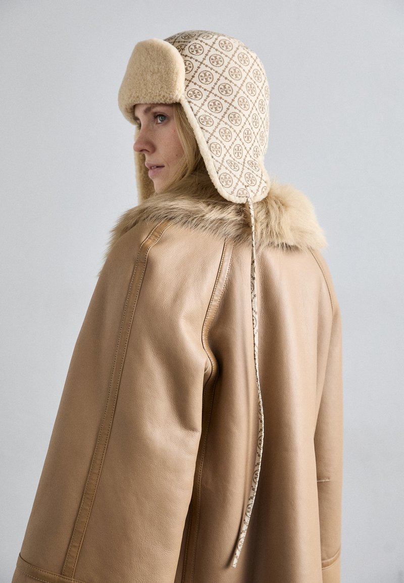 Beige leather coat with fur trim and a patterned, cream hat. The hat features ear flaps and a textured design, showcasing intricate details.