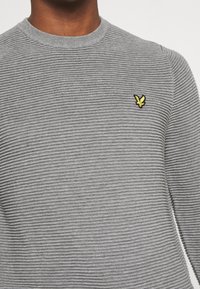 Grå ribstrikket sweater med rund halsudskæring og en lille gul fuglelogo på brystet. Stoffet virker blødt og tekstureret.