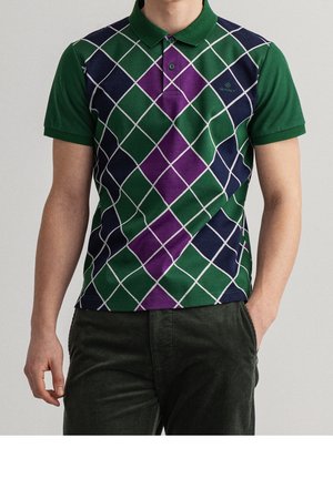 Homme portant un polo à manches courtes avec un motif losanges vert, violet et bleu marine et un pantalon vert foncé, debout avec une main dans la poche.