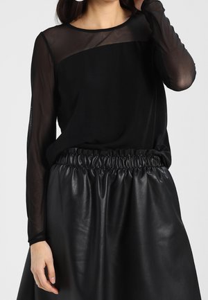 Woman wearing a black sheer long-sleeve top with a solid panel and a high-waisted black leather skirt with gathered waistband.