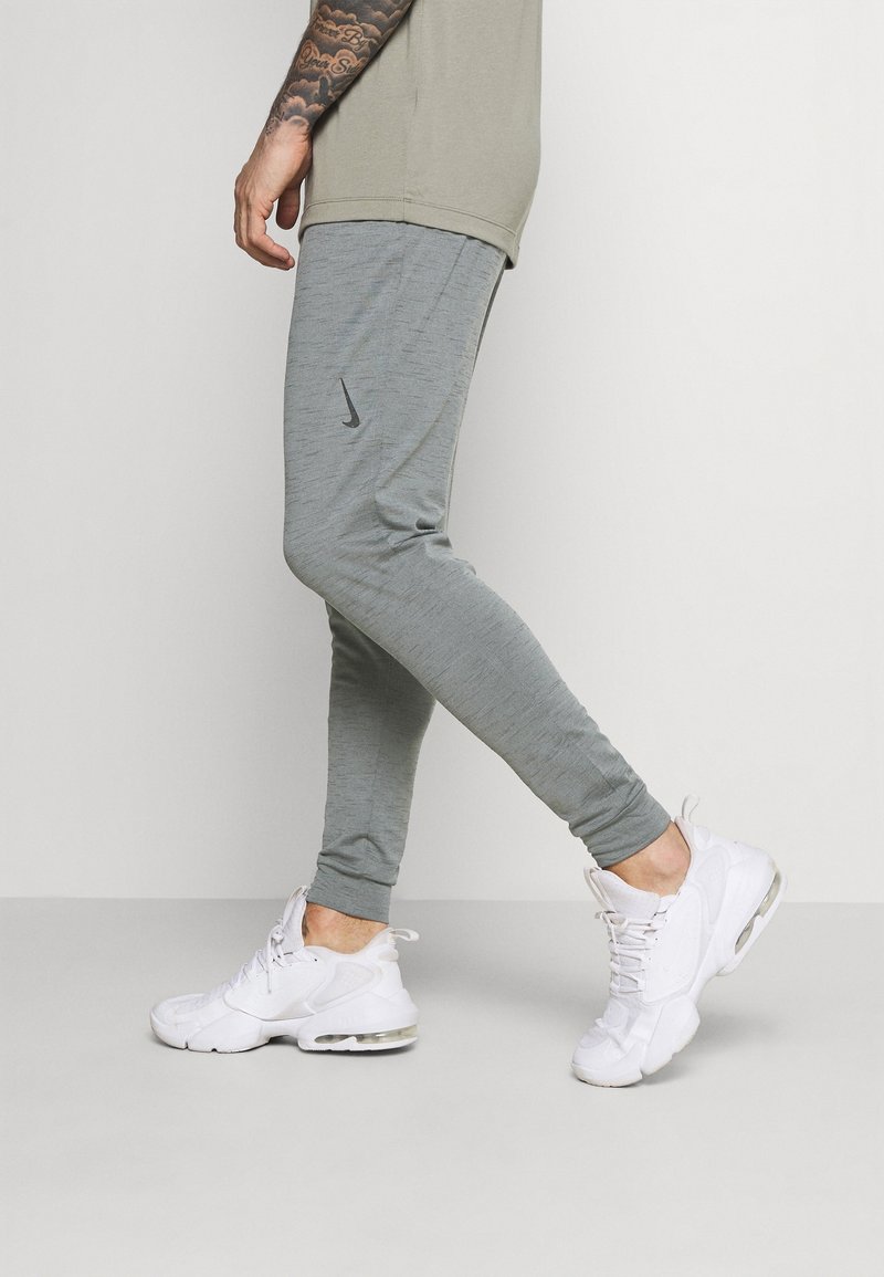 Muž v šedých teplácích Nike a bílých teniskách jde, ukazuje tetovanou ruku a béžové tričko na jednobarevném bílém pozadí.