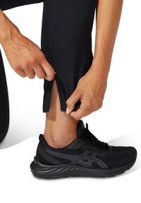 Zwarte sportbroek met een ritssluiting aan de onderkant, gecombineerd met zwarte hardloopschoenen met een bovenwerk van mesh en een gevoerde zool.