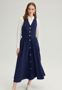 Robe navy avec un haut ajusté en gilet et une jupe pleine et fluide. Comprend des boutons dorés sur le devant et une chemise à col blanc en dessous.