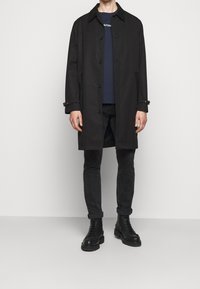 Black trench coat with a collar and button closures, worn over a navy shirt. Paired with black jeans and chunky black combat boots.
