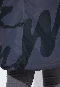 Giacca impermeabile di colore grigio scuro con una finitura lucida. Presenta motivi astratti neri. Dettaglio delle cuciture visibile all'orlo.