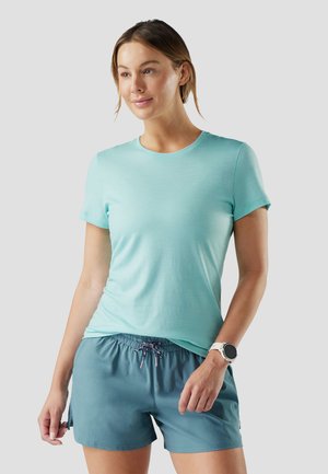 Kobieta w jasnoniebieskiej koszuli z krótkim rękawem i turkusowych szortach wiązanych na sznurek, z białym smartwatchem na lewej ręce, stoi na tle szarego tła.