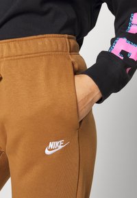 Bruna sweatpants med mjuk textur, med sidofickor och en vit Nike-logotyp på låret. Midjan är elastisk.