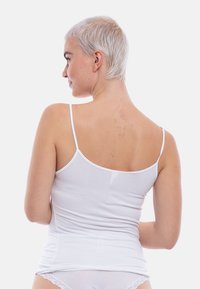 Giovane con capelli platino biondi corti, che indossa un canottiera bianca e culotte in pizzo bianco, visto di spalle, su uno sfondo semplice.
