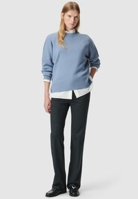 Lichtblauwe gebreide trui met ribbels aan de onderkant en mouwen, gedragen over een witte blouse met kraag, gecombineerd met donkergrijze broeken en zwarte schoenen.