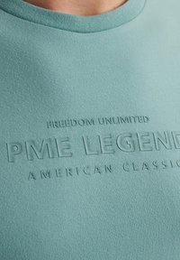 Majica iz mehkega, teal materiala z rebrastim okroglim izrezom. Ima embosirano besedilo: "SVOBODA NEOMEJENA PME LEGEND AMERIKANSKA KLASIKA."