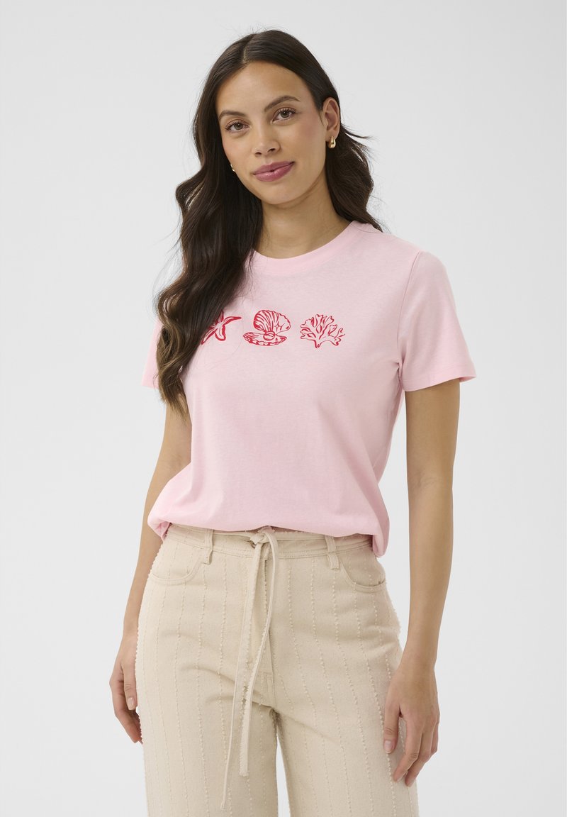 Giovane donna con lunghi capelli scuri che indossa una maglietta rosa con disegni di conchiglie rosse e pantaloni beige con coulisse, in piedi contro uno sfondo bianco.
