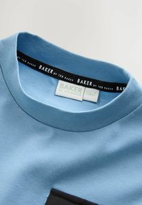 Svetlo modré tričko s okrúhlym výstrihom a čiernym lemom, na štítku je nápis "BAKER by Ted Baker" a biely štítok veľkosti s textom "Vyrobené v Turecku."
