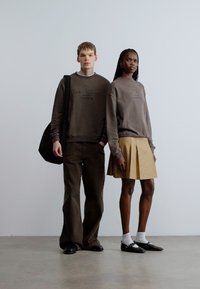 Dos modelos están sobre un fondo liso. Uno lleva un suéter y pantalones marrones; el otro lleva un suéter gris y una falda amarilla a rayas.