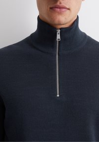 Mörkblå halvdraget tröja med ribbad textur, hög krage och silverfärgad dragkedja. Tyget har ett mjukt, stickat utseende.