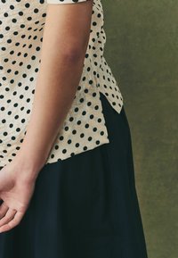 Polkadot skjorte i cremefarvet med sorte prikker, korte ærmer og splittede kantdetaljer, parret med en mørk nederdel; glat tekstur synlig.