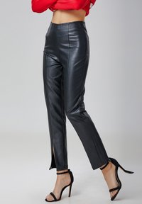 Pantalons en simili cuir noir à coupe slim, avec des fentes sur les côtés au niveau des chevilles et une taille haute. Portés avec des talons aiguilles noirs.