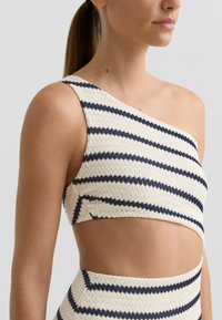 Aszimmetrikus, egyrészes fürdőruha felsőrész krémszínben, tengerészkék csíkokkal. Texturált anyag, kivágott dizájn, bordázott mintával.