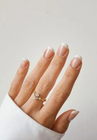 Złoty pierścionek z trzema diamentopodobnymi akcentami, założony na ręce z naturalnie wyglądającymi paznokciami w stylu francuskim.