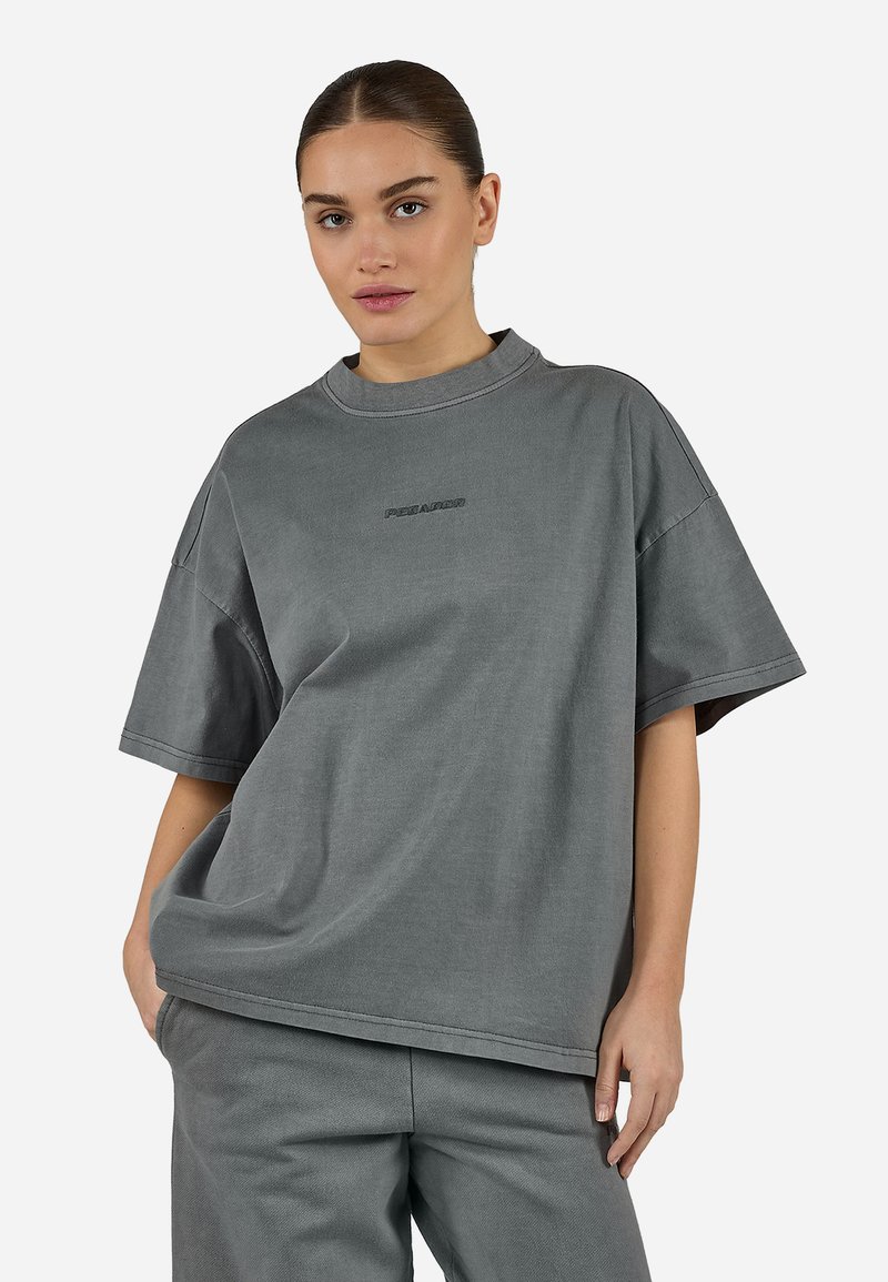 Νεαρή γυναίκα που φοράει χαλαρό γκρι κοντομάνικο πουκάμισο και ταιριαστό γκρι παντελόνι, στέκεται με το ένα χέρι στην τσέπη μπροστά σε ένα απλό φόντο.