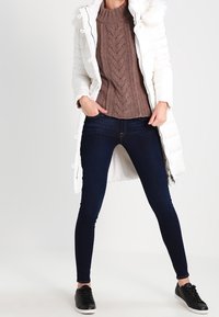 Abrigo de plumas blanco con capucha de piel sintética, suéter de punto trenzado marrón, jeans ajustados oscuros y zapatillas negras. Conjunto casual en capas.