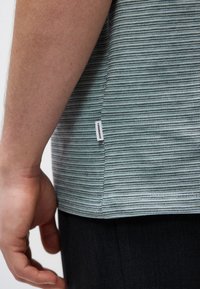 Κοντινό πλάνο του χεριού και του βραχίονα ενός ατόμου δίπλα σε πουκάμισο με ρίγες σε ανοιχτό μπλε και γκρι χρώμα, με μια μικρή υφασμάτινη ετικέτα "Lindbergh" στη πλευρική ραφή.