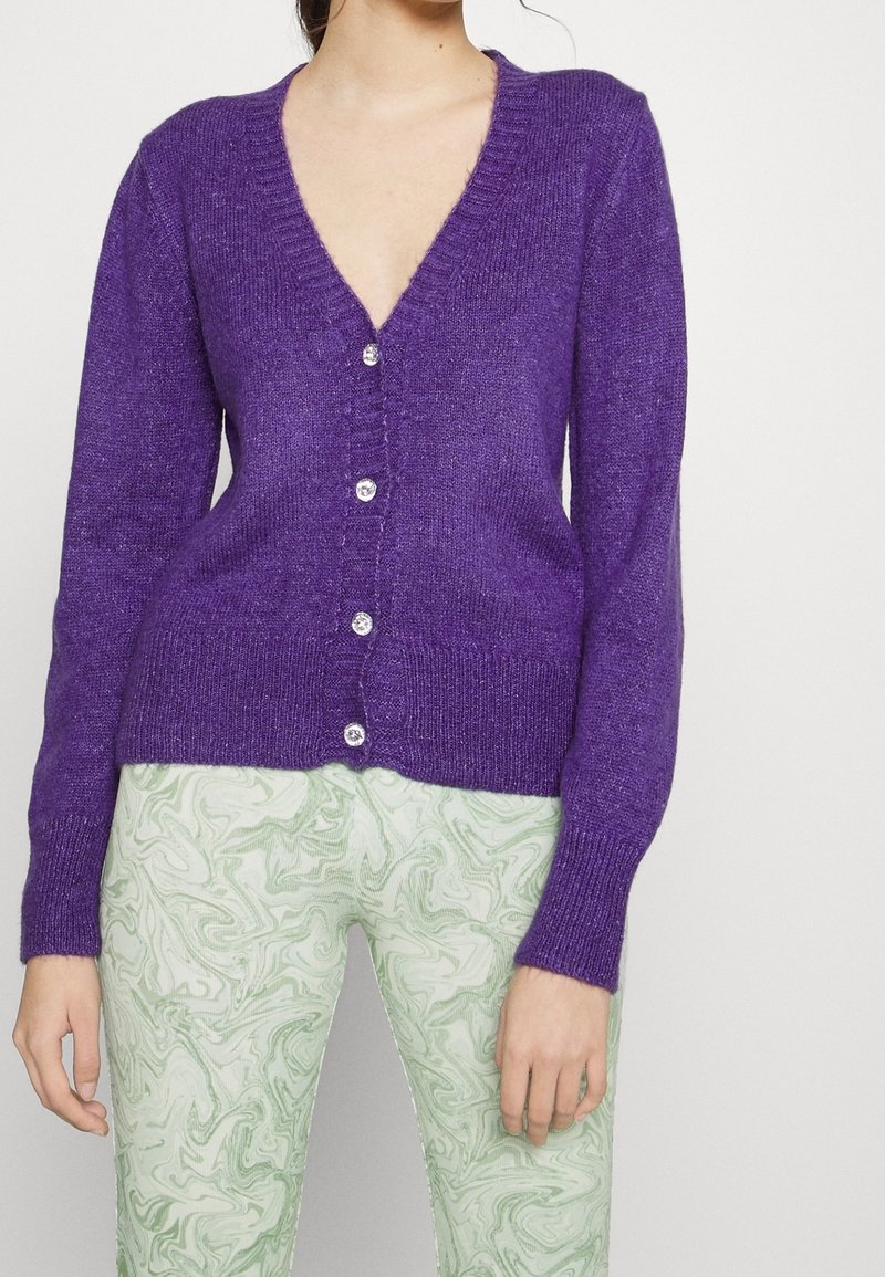 Lila cardigan med V-hals, långa ärmar och fem genomskinliga knappar framtill. Tyget verkar mjukt och texturerat. Matchas med gröna marmorerade byxor.