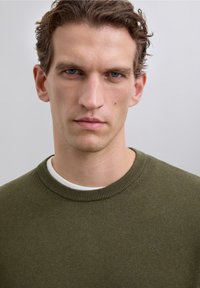 Oliwkowo zielony sweter z okrągłym dekoltem, prążkowaną teksturą i subtelnym logo na piersi, noszony na białej koszuli.