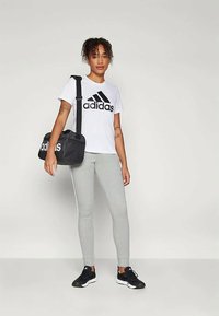 Γυναίκα φορώντας άσπρη μπλούζα Adidas, γκρι κολάν και μαύρα sneakers, κρατώντας μια μαύρη τσάντα Adidas στον ώμο της, να στέκεται μπροστά σε έναν απλό τοίχο.