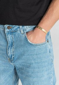 Jeans de mezclilla azul claro con un corte clásico, que presentan costuras visibles y un bolsillo lateral. Se lleva una pulsera dorada en la muñeca.
