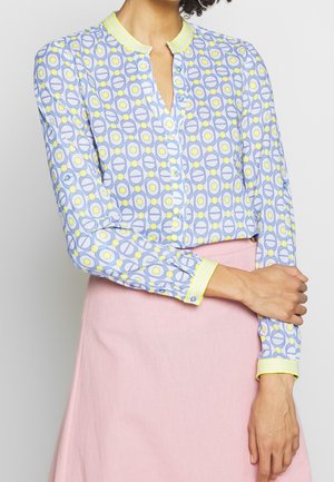 Vrouw draagt een blouse met lange mouwen en knopen met een blauw, geel en wit geometrisch patroon en een lichtroze rok met hoge taille.