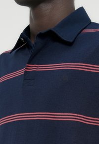 Κοντινό πλάνο ενός ατόμου που φοράει μπλε πουκάμισο πόλο με κόκκινες οριζόντιες ρίγες και γιακά, σε ουδέτερο φωτεινό φόντο.