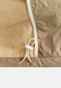 Tessuto impermeabile beige con finitura strutturata, dotato di un toggle con cordino bianco e cuciture a contrasto lungo i bordi.