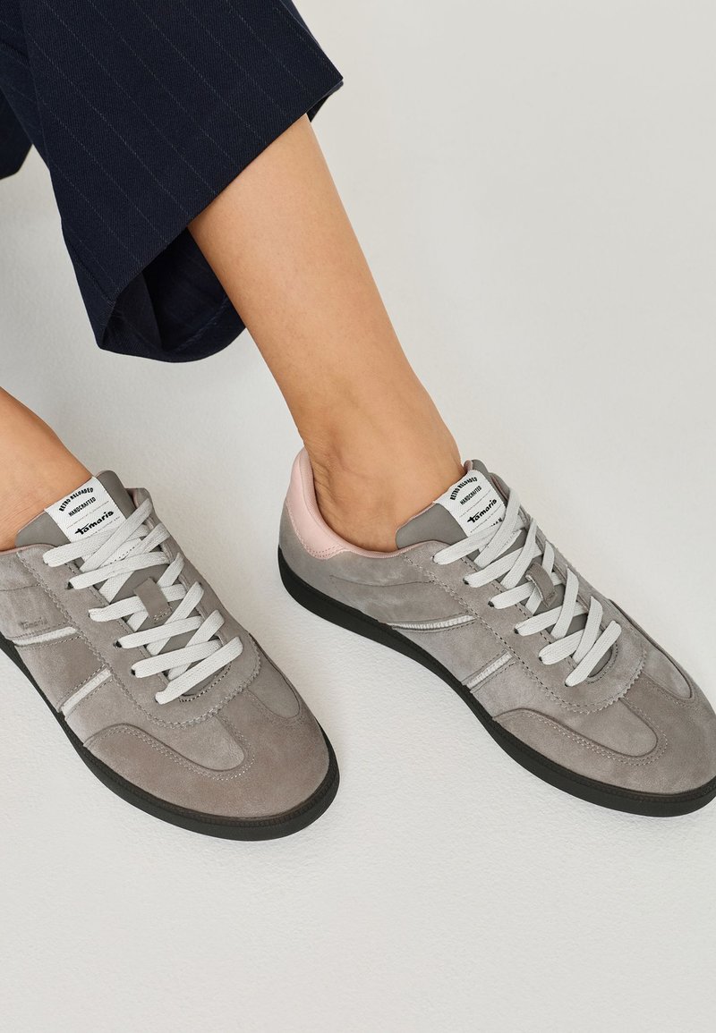 Zapatillas de ante gris con cordones blancos y suelas negras. Con un toque de rosa claro en el talón, presentan un diseño atlético clásico.