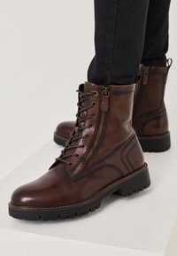 Bottes en cuir marron avec un bout rond, des lacets à l'avant, une fermeture éclair sur le côté et une semelle en caoutchouc noir robuste avec un motif de traction.