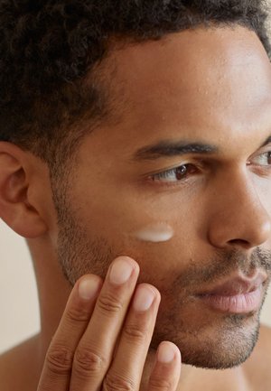 Feuchtigkeitsspendende Creme auf der Wange eines Mannes aufgetragen, die eine glatte, cremige Textur zeigt. Die Haut ist mitteltonig mit Stoppeln, die Finger berühren sanft.