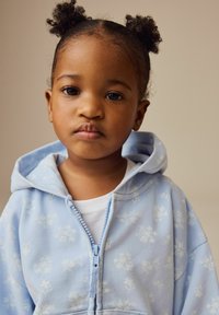 Lyseblå zip-up hoodie med blomstermønster, blød tekstur, hætte med snor, båret over en hvid T-shirt. Barn med to knoldestile.