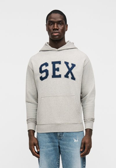 Pilkas džemperis su gobtuvu, priekyje mėlyna siuvinėta užrašas „SEX“, su kengūros tipo kišene ir ribotais rankogaliais. Dėvimas su šviesiai mėlynais, nudraskytais džinsais.