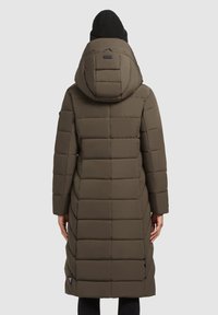 Gewatteerde olijfgroene jas met een capuchon, voorzien van een gequilt ontwerp, lange mouwen en zijritsen, gedragen over zwarte broeken en een gebreide muts.